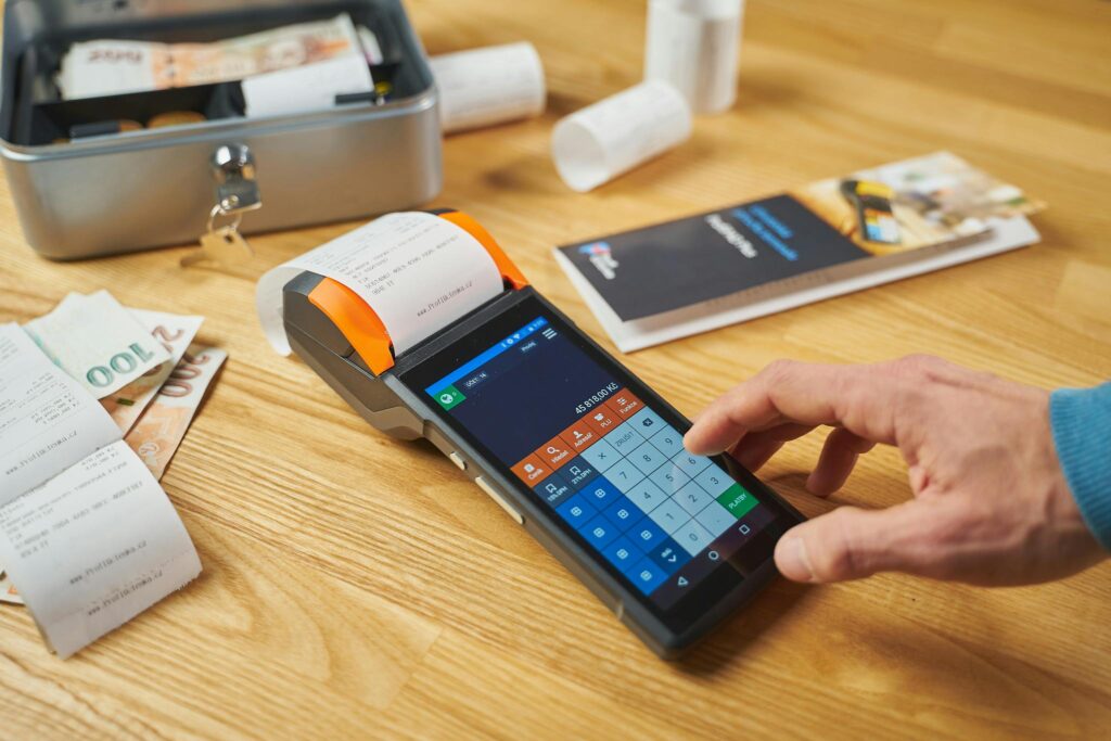 Person using a digital payment terminal on a wooden desk with cash and receipts visible.